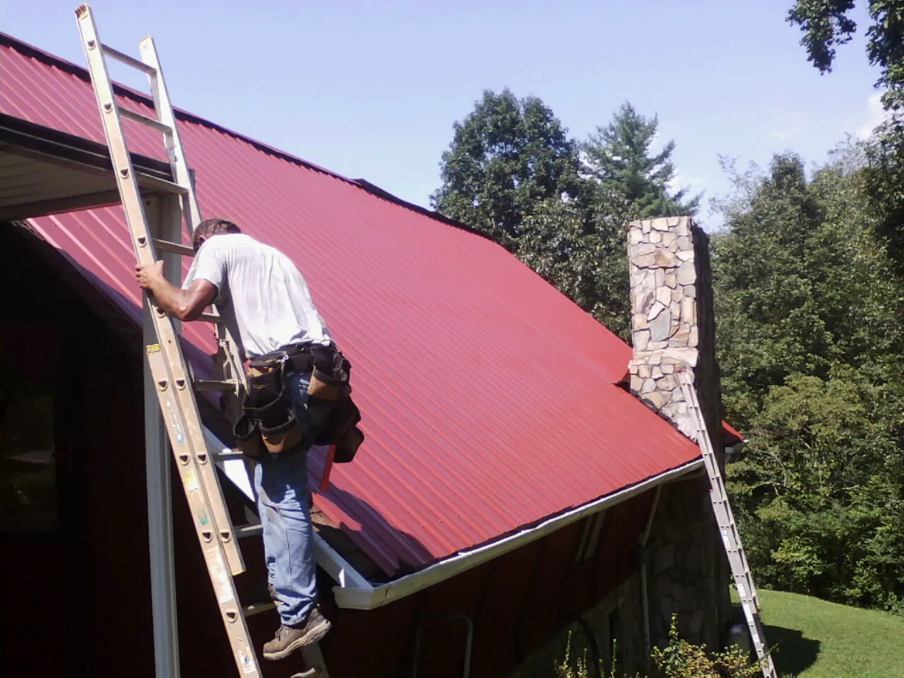 Roofer on a ladder beside a red metal roof and stone chimney — Soffit & Fascia Repair by Roofing Brothers in Roseland